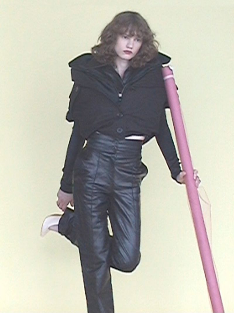 Model wearing a black puffy vest and leather trousers leaning against a tall pink prop against a plain background