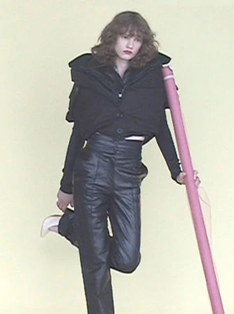 Model wearing a black puffy vest and leather trousers leaning against a tall pink prop against a plain background
