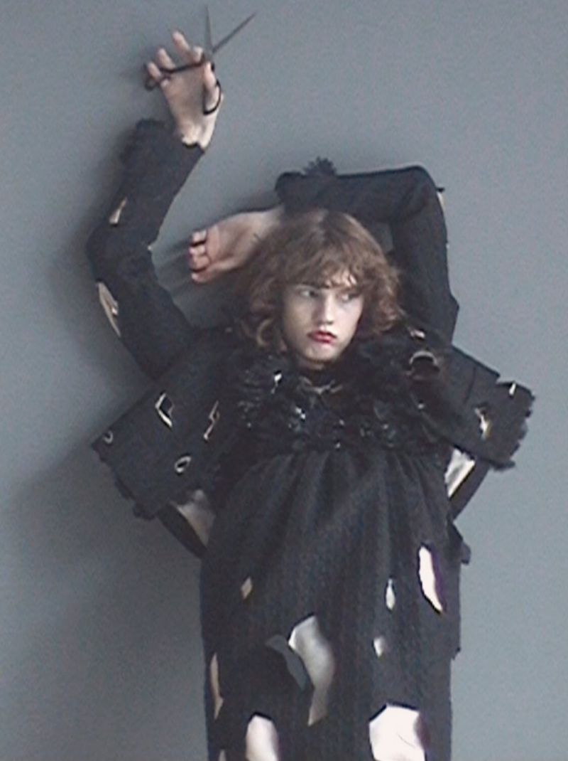 Model wearing a distressed cut-out black garment holding scissors overhead against a plain grey studio backdrop.