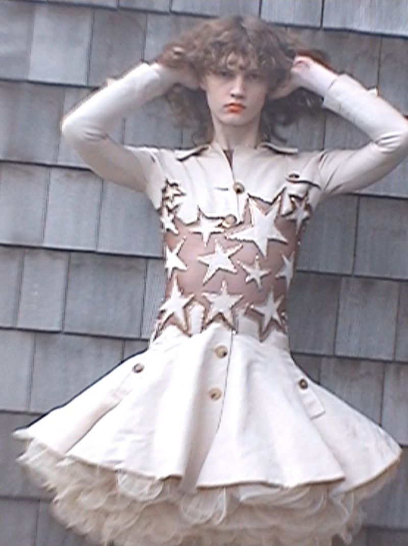 Model with wavy brown hair posing with hands on head wearing a cream star-embellished flare dress against a wooden shingle wall.