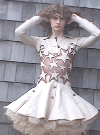 Model with wavy brown hair posing with hands on head wearing a cream star-embellished flare dress against a wooden shingle wall.