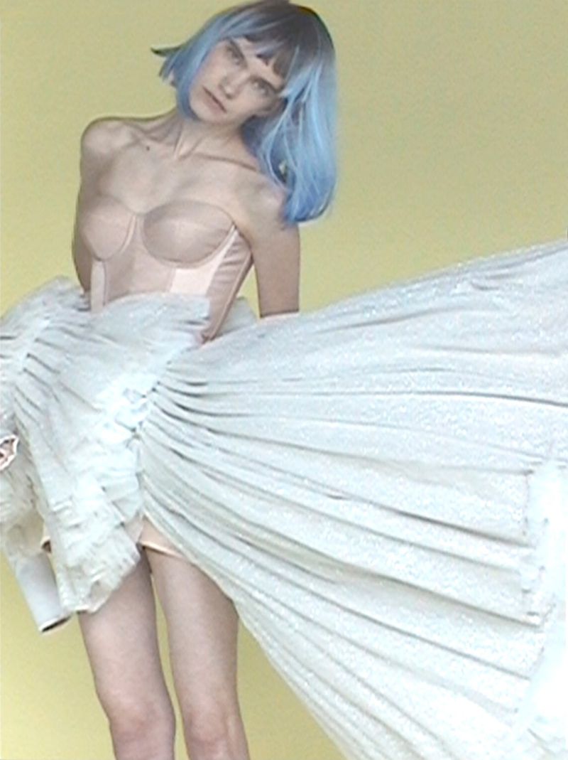 Model with blue hair in a blush corset top and an expansive pleated white skirt against a pale yellow background.