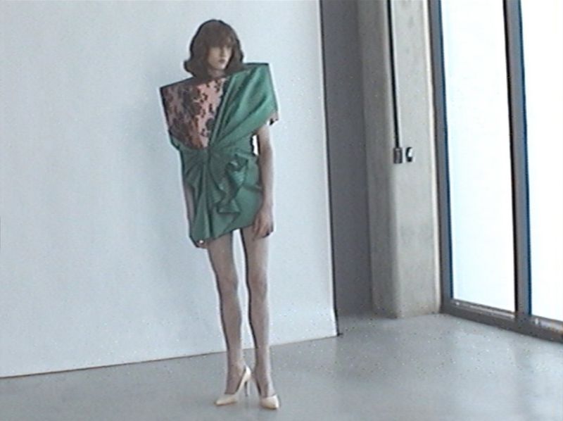 Model posing in an avant-garde green structured mini dress with pointed high heels beside a studio window.