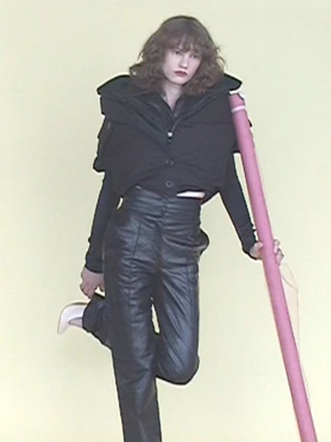 Model wearing a black puffy vest and leather trousers leaning against a tall pink prop against a plain background