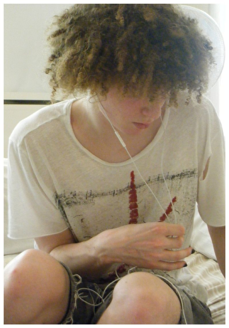 Seated person with voluminous curly hair wearing a white graphic t-shirt and white wired earphones in a bright bedroom.