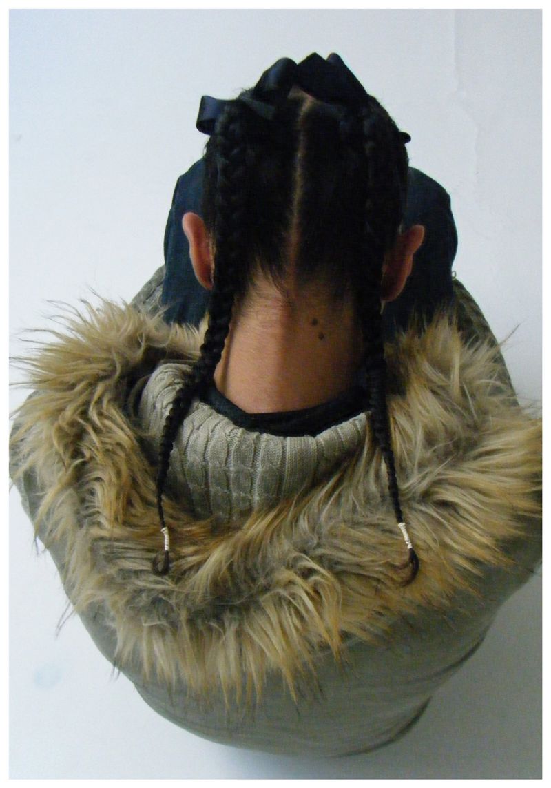 High-angle back view of braided pigtails with black ribbons, wearing a fur-trimmed hooded parka against a white studio floor.