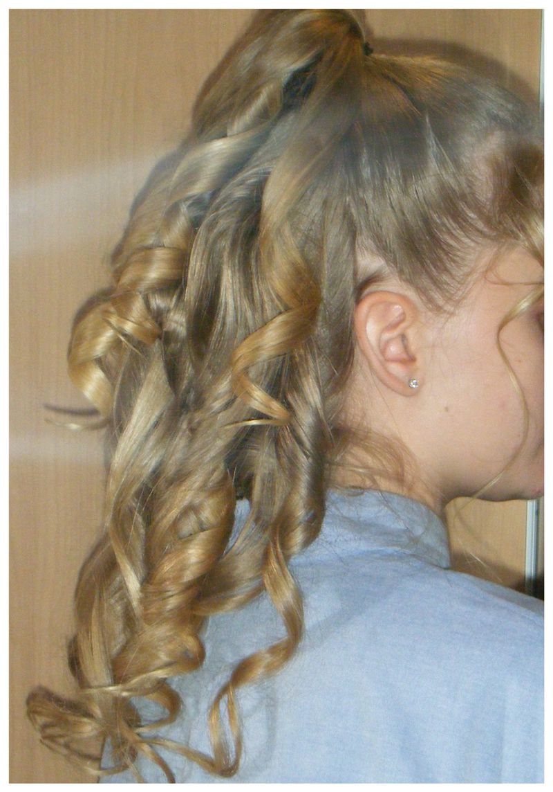 Side-back view of a woman with blonde hair styled into a high curly half-up ponytail wearing a light blue top.