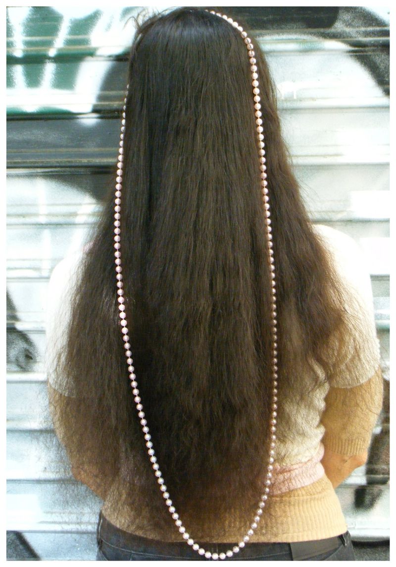 Back view of a model with very long brown hair draped from crown to waist with an elongated strand of pearls.