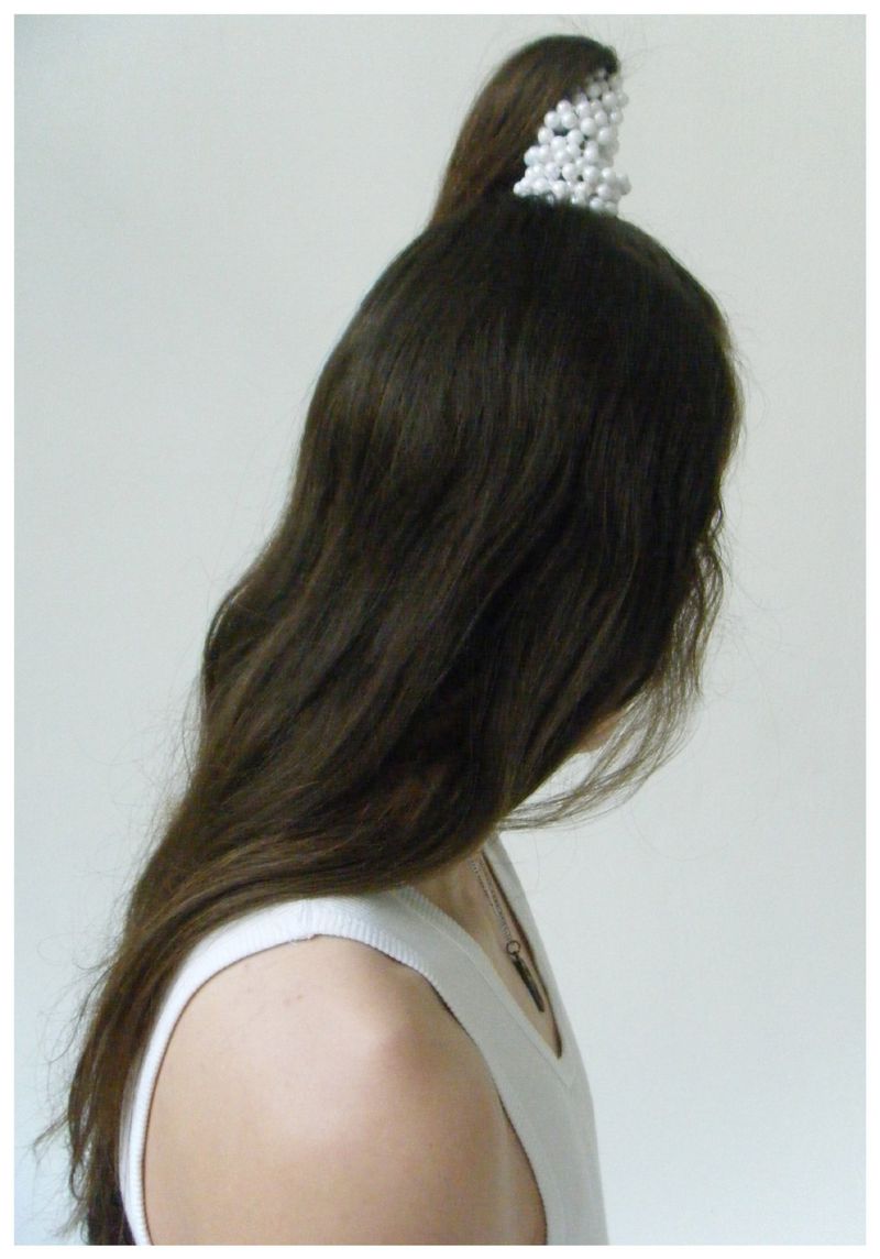 Model facing away with long brown hair and a sculptural white pearl headpiece, wearing a white tank top against a plain studio background.