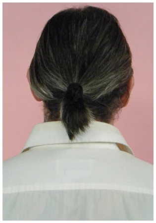 Rear view of a person with dark grey-streaked hair tied into a low ponytail wearing a white collared shirt against a pink studio backdrop.