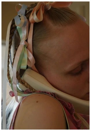 Close-up of a blonde woman resting her head sideways against a cream handset, wearing braided hair adorned with colorful pastel ribbons.