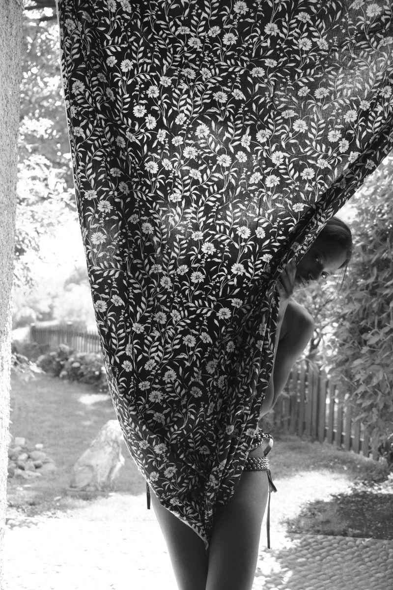 Model Wendy Garcia partially hidden behind a draped floral print fabric outdoors in a sunlit garden setting in black and white.