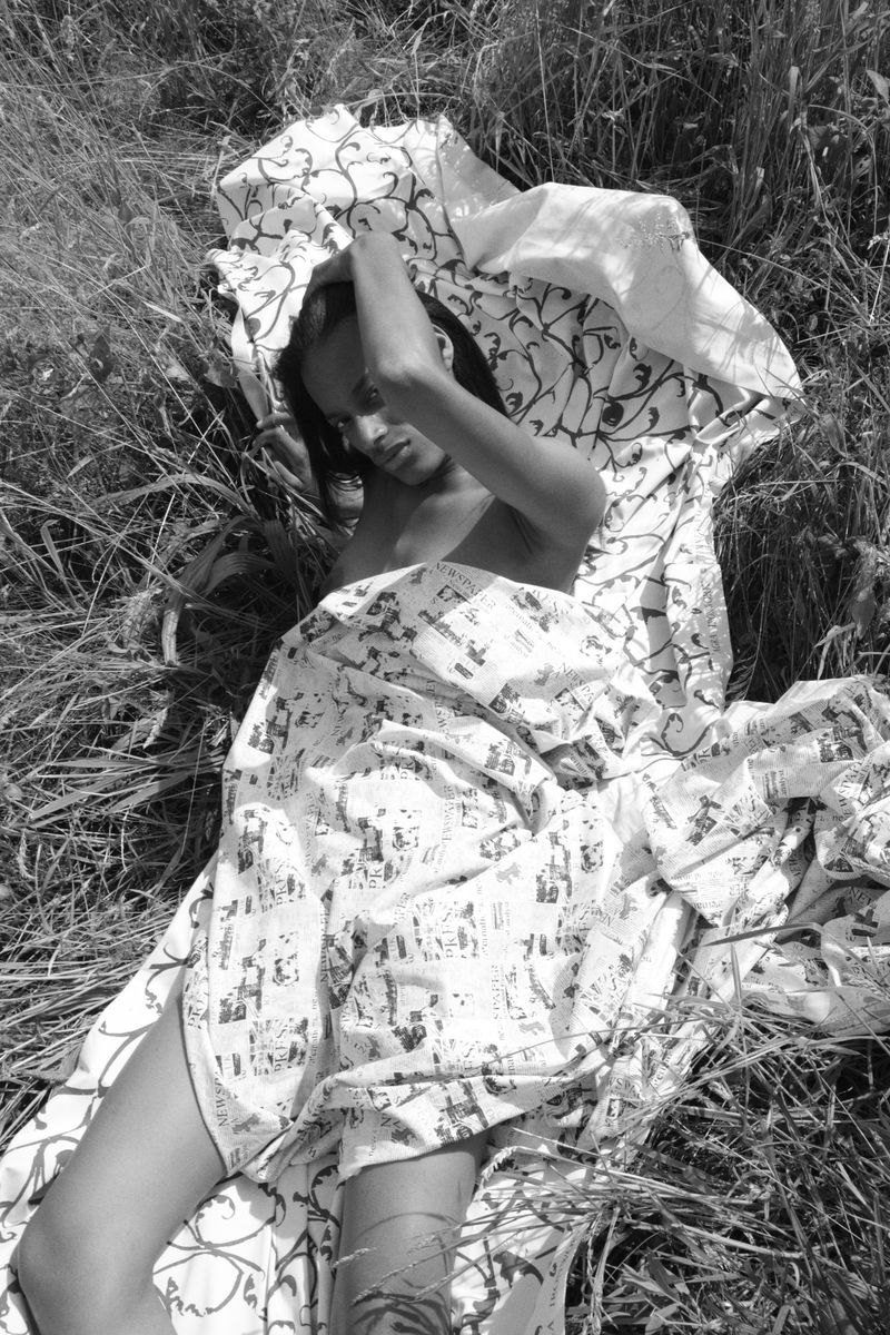 Model lying amongst tall grass wrapped in a patterned white fabric sheet