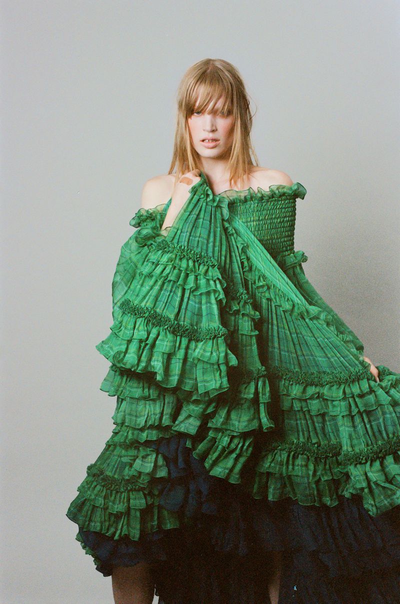 Model Jacqui Hooper posing in a voluminous tiered green ruffle dress against a pale neutral studio backdrop.