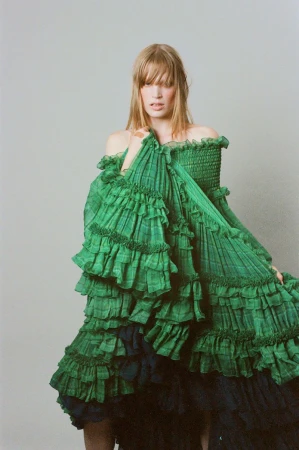 Model Jacqui Hooper posing in a voluminous tiered green ruffle dress against a pale neutral studio backdrop.