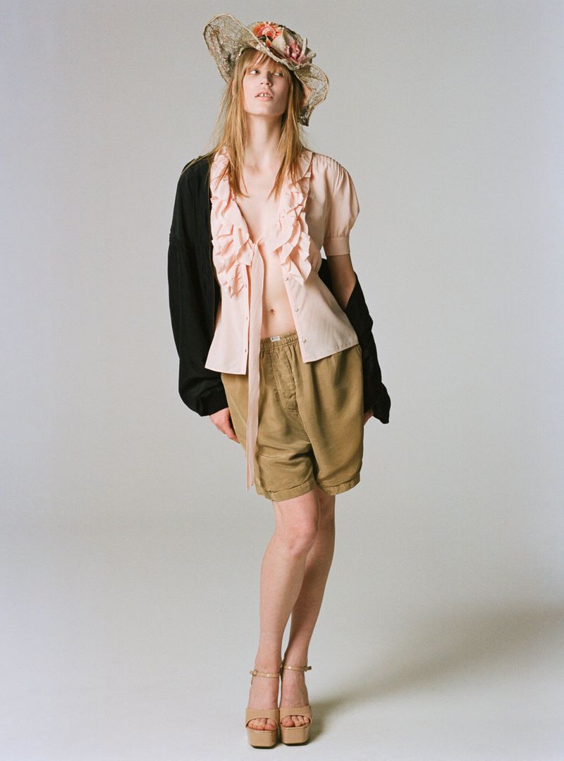 Model Jacqui Hooper standing in a ruffled pink top, khaki shorts, black cardigan, and straw floral hat against a white studio background.