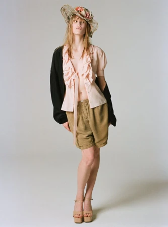 Model Jacqui Hooper standing in a ruffled pink top, khaki shorts, black cardigan, and straw floral hat against a white studio background.