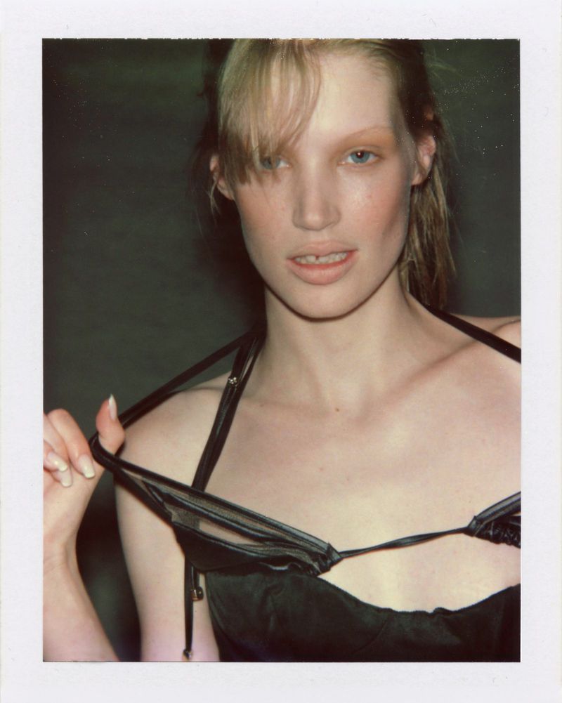 Polaroid portrait of model Jacqui Hooper pulling straps of a black satin top against a dark studio background.