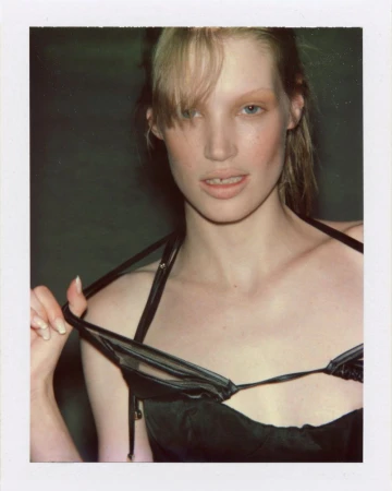 Polaroid portrait of model Jacqui Hooper pulling straps of a black satin top against a dark studio background.