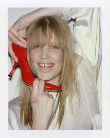 Model Jacqui Hooper smiles while holding a red high-heeled shoe up to her face in a Polaroid-style portrait against a white backdrop.