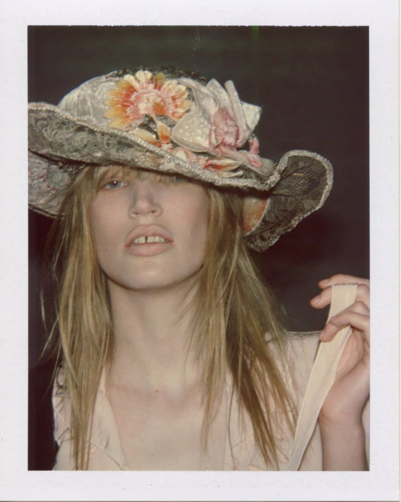 Model Jacqui Hooper wearing an ornate floral textured hat and a pale ruffled garment in a Polaroid portrait.