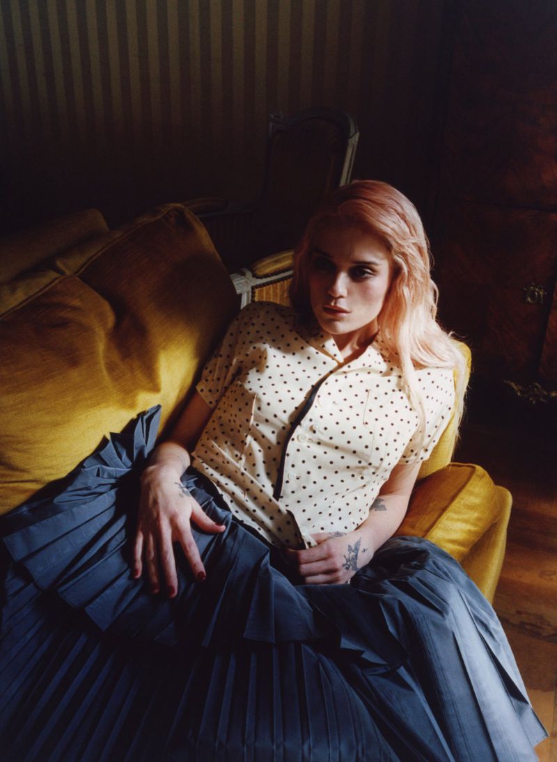 Sky Ferreira wearing a cream polka dot shirt and dark pleated skirt reclining on a yellow velvet chair.