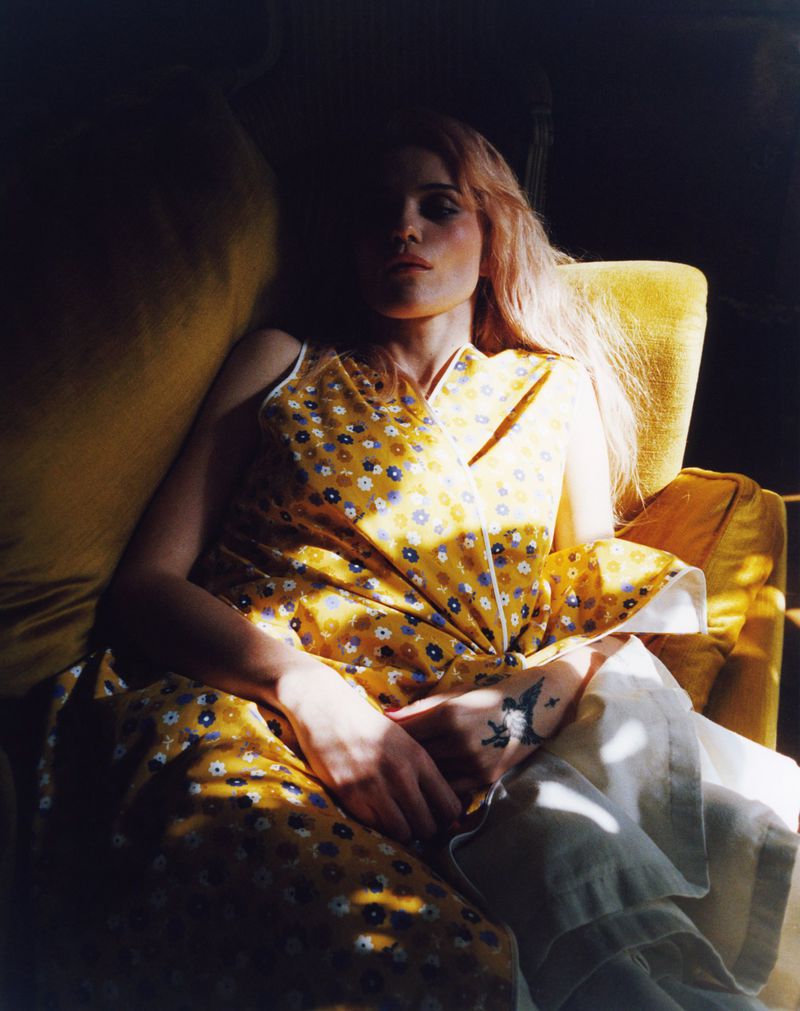 Sky Ferreira wearing a yellow floral top reclining in a yellow velvet armchair in dappled sunlight.
