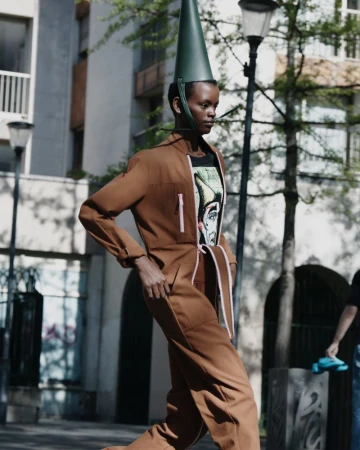 A fashion-forward adult female strides outdoors in a brown jumpsuit with a conical hat. Building and trees form the background.