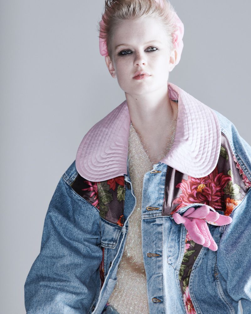 A young woman poses in a studio, wearing a denim jacket with floral accents and a light pink hat, complemented by a silver top.