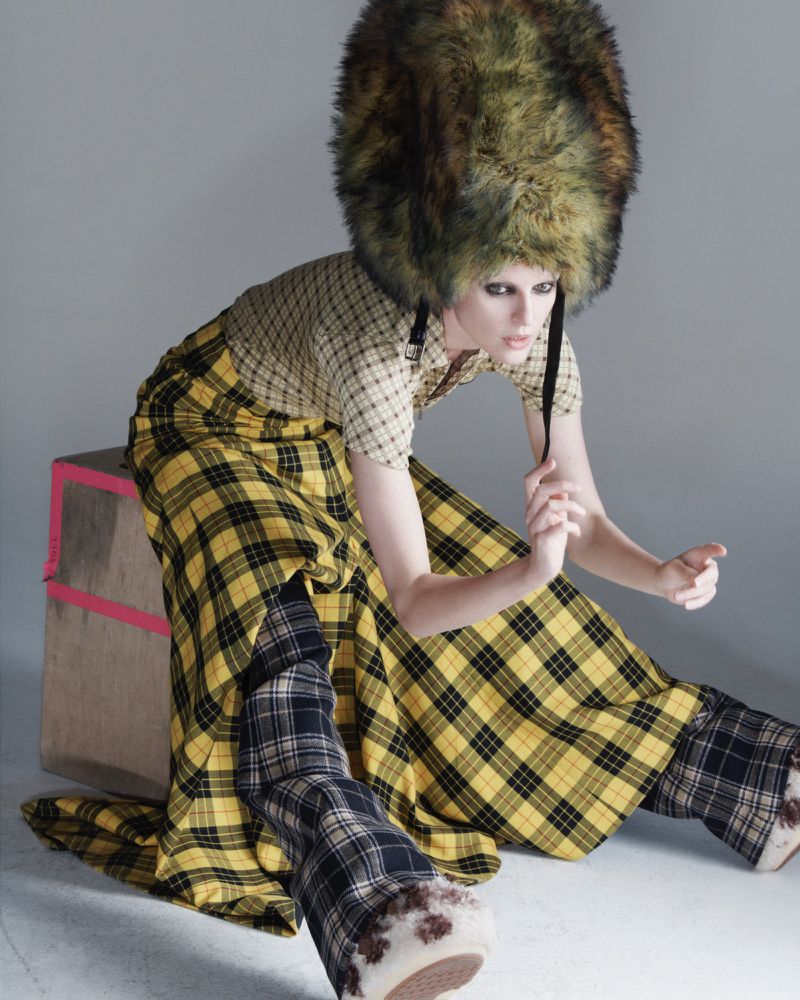 Fashion model wearing an oversized tall fur hat and yellow plaid skirt posed beside a wooden box against a neutral studio backdrop.