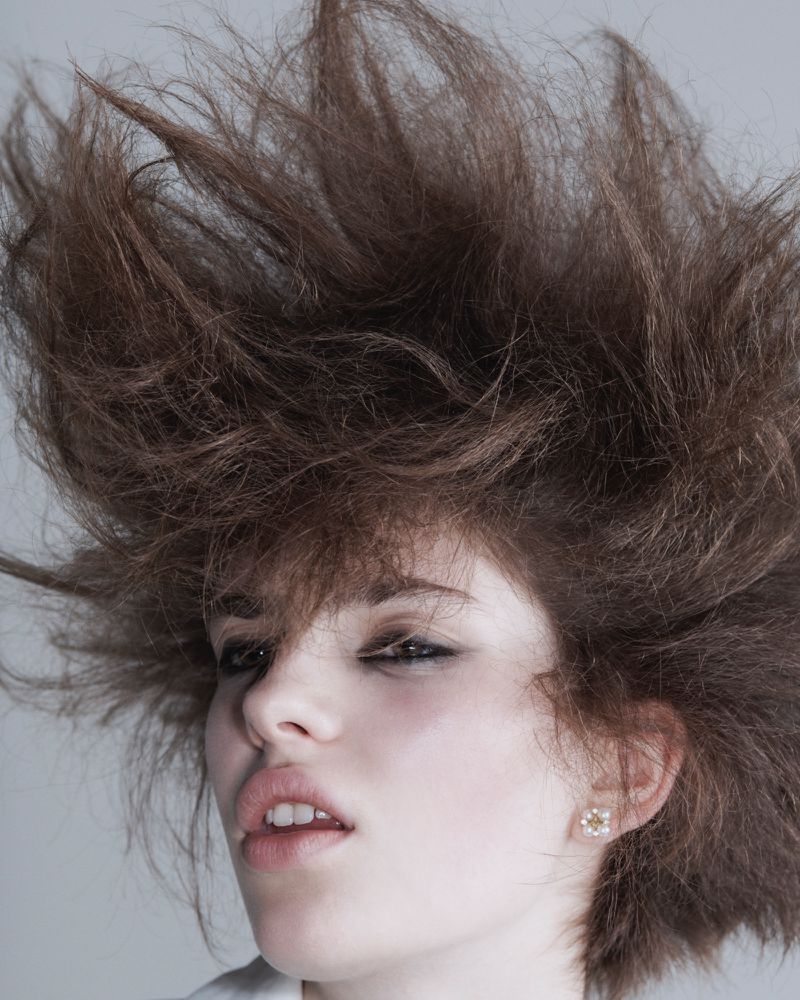 Close-up portrait of a model with voluminous teased brown hair, smoky eye makeup, and a small stud earring against a pale backdrop.