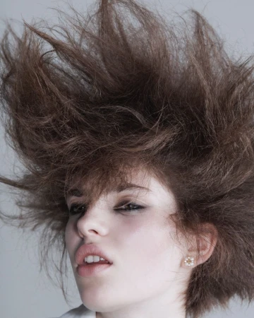 Close-up portrait of a model with voluminous teased brown hair, smoky eye makeup, and a small stud earring against a pale backdrop.