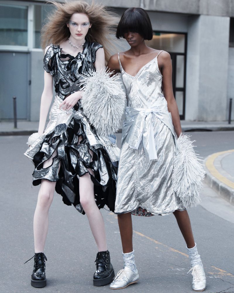 Two fashion models in silver metallic dresses pose outdoors. One model with fair skin and the other with dark skin stand close.