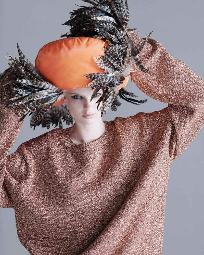 Woman in a fashionable orange hat adorned with feathers and wearing a shimmering bronze sweater, posing against a neutral grey backdrop.