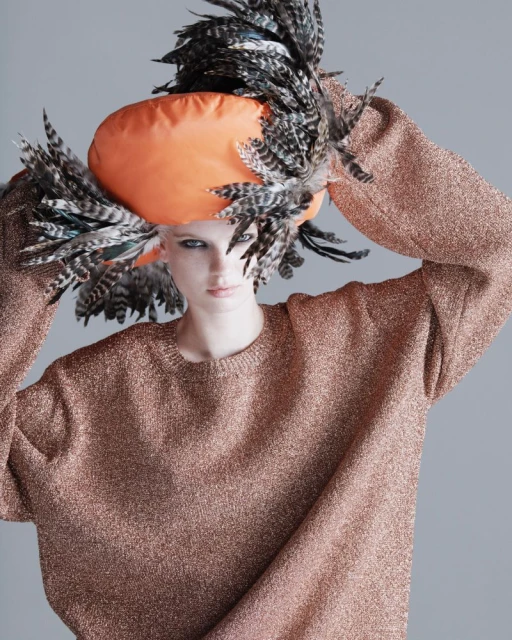 Woman in a fashionable orange hat adorned with feathers and wearing a shimmering bronze sweater, posing against a neutral grey backdrop.