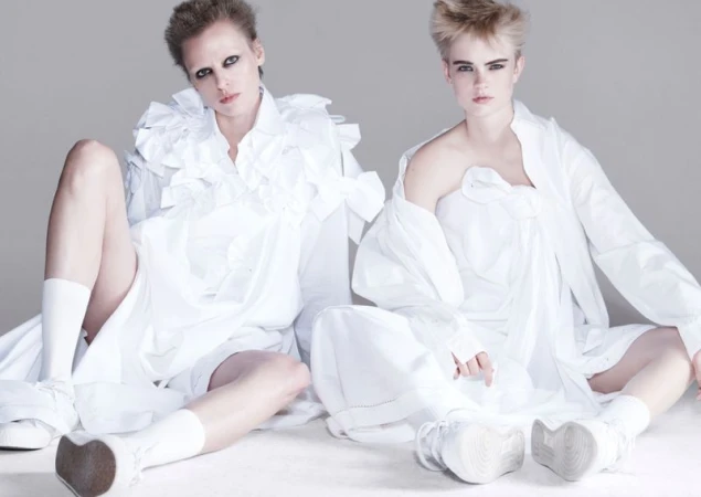 Two fair-skinned women in white shirts and sneakers pose against a grey background in a fashion studio, showcasing edgy editorial style.