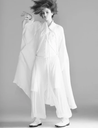 Monochromatic fashion shot: a young woman in white harem pants, tunic, and boots poses in a photo studio with a neutral gray background.