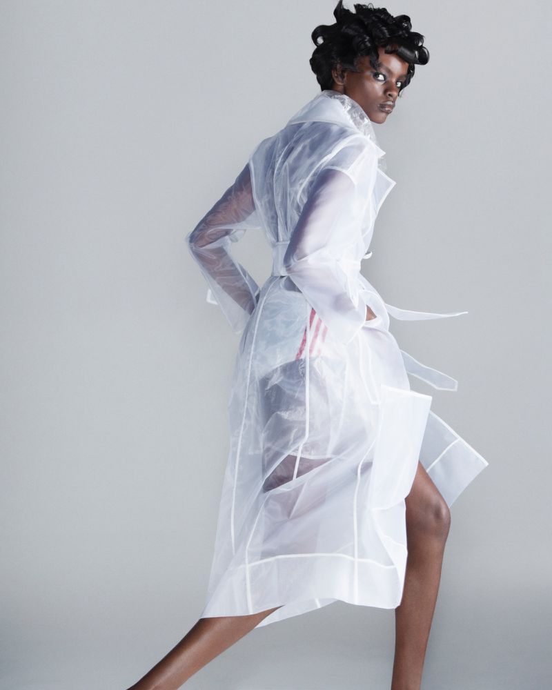 Fashion shot of a Black woman in a sheer white trench coat with a tie waist, standing against a neutral gray background in a photo studio.
