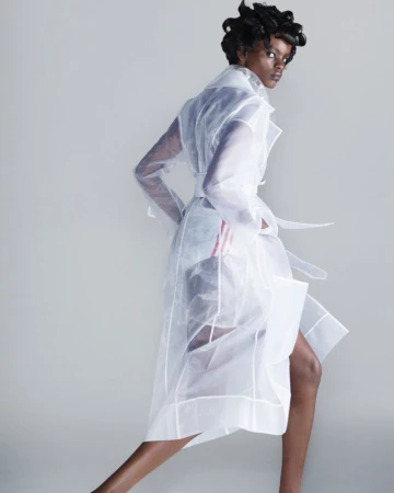 Fashion shot of a Black woman in a sheer white trench coat with a tie waist, standing against a neutral gray background in a photo studio.