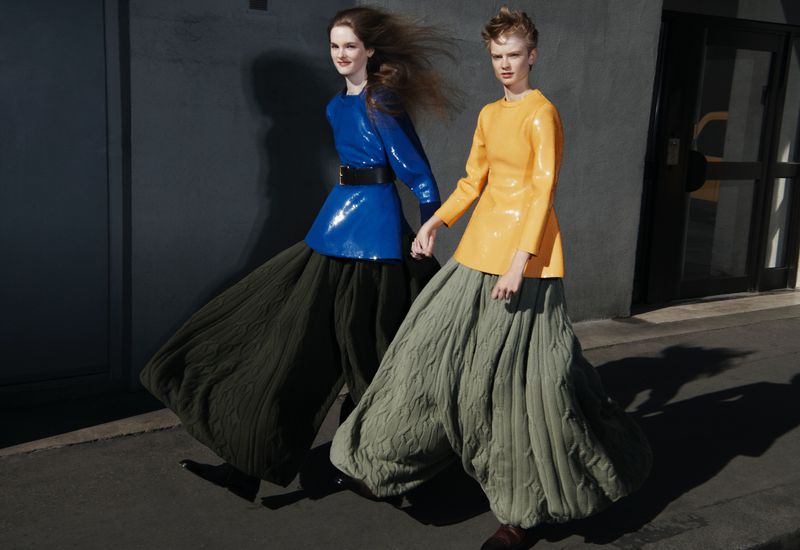 Two women in yellow and blue tops, hold hands in an outdoor fashion shoot. They wear billowing, cable-knit trousers, and face the camera.