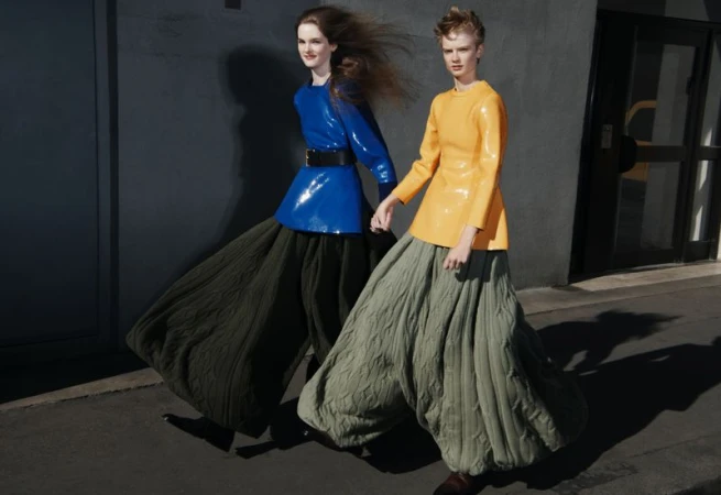 Two women in yellow and blue tops, hold hands in an outdoor fashion shoot. They wear billowing, cable-knit trousers, and face the camera.