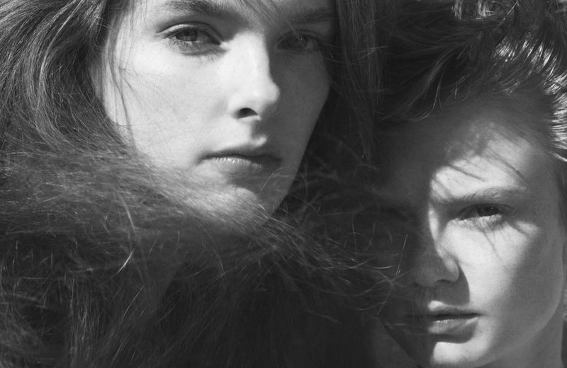 Black and white portrait of two young women with long hair, one in sharp focus, the other with soft shadows and dramatic lighting.