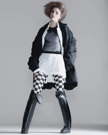 Fashionable woman in oversized black coat poses in a photo studio with grey backdrop. She wears checkered leggings and black boots.