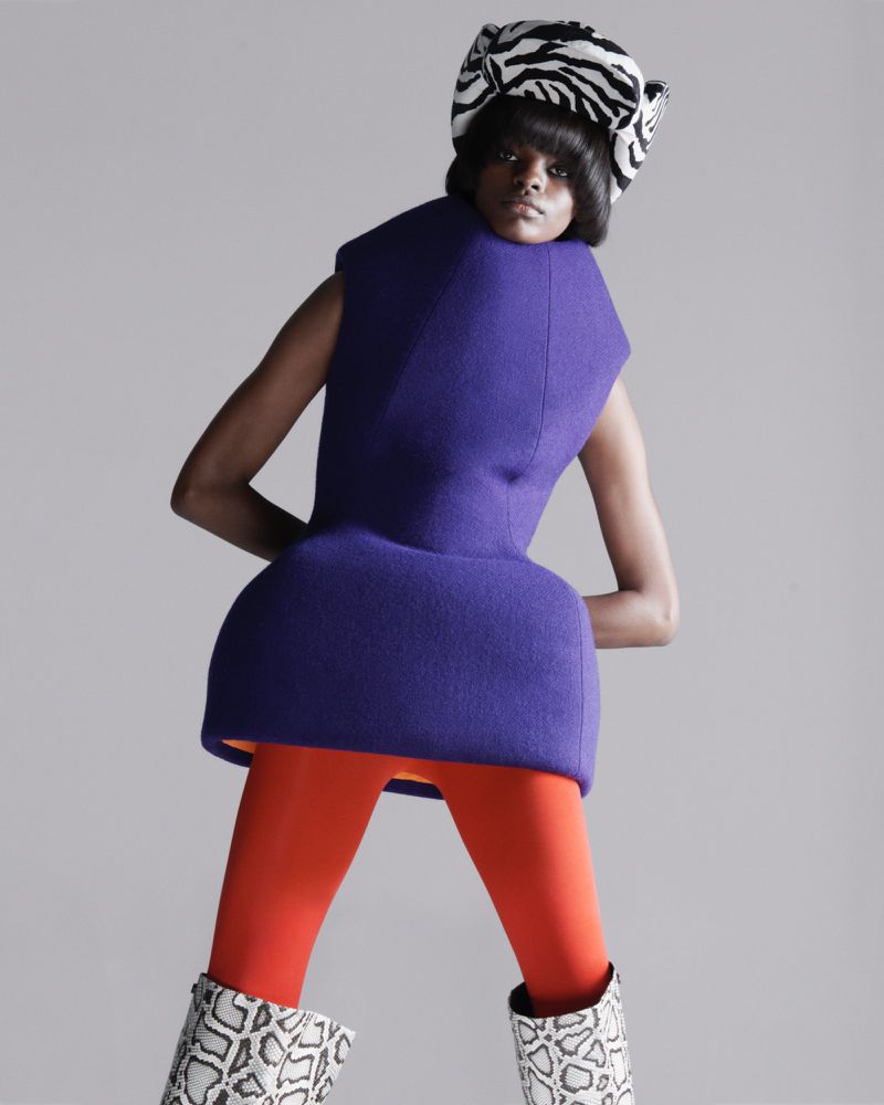 A fashion portrait of a woman wearing a zebra-striped turban, a purple dress, orange leggings, and snakeskin boots in a photo studio.