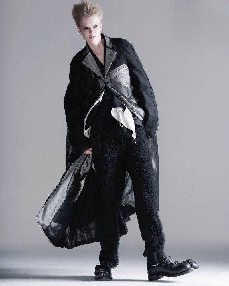 Fashion editorial shot of a female model with a modern, short blonde hairstyle wearing a unique layered black and grey outfit.