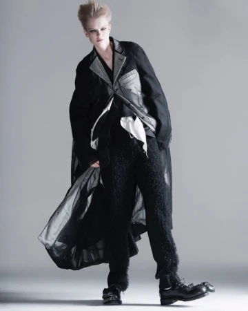 Fashion editorial shot of a female model with a modern, short blonde hairstyle wearing a unique layered black and grey outfit.