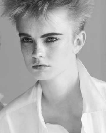 Black and white close-up portrait of a model with short spiky hair and freckles wearing an open white collared shirt.