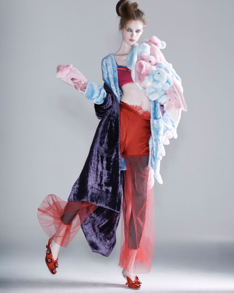 Fashionable woman in an avant-garde outfit, including a long coat, red shorts, tulle pants, and heels, posing against a grey backdrop.