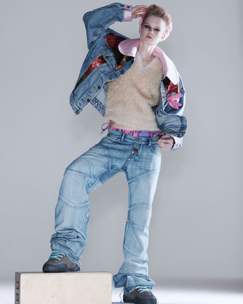 Fashion model in denim jacket and jeans poses on a box in a studio. The subject showcases a unique and stylish denim outfit in neutral setting.