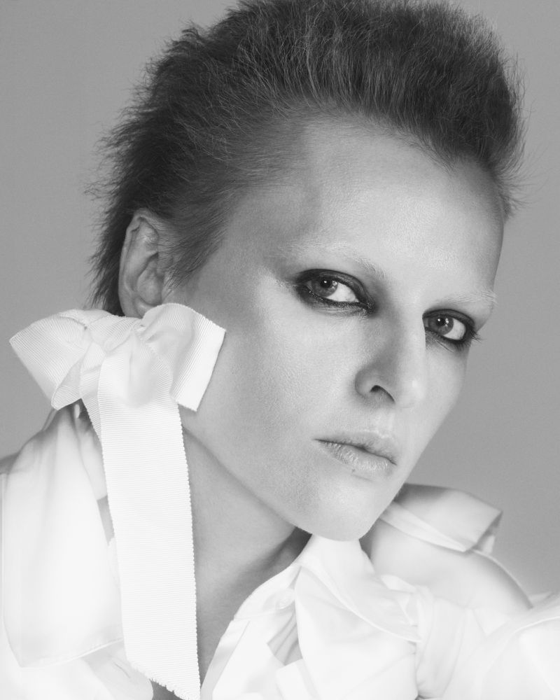 Monochrome portrait of a person with short hair and pale skin wearing a white collared shirt with a large satin ribbon at the neck.